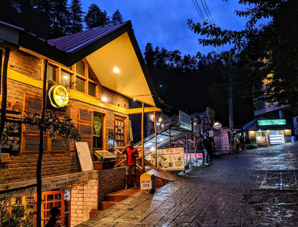 Scenic commercial street in Manali featuring hotels, shopping stores, mountain town architecture, and visitors.