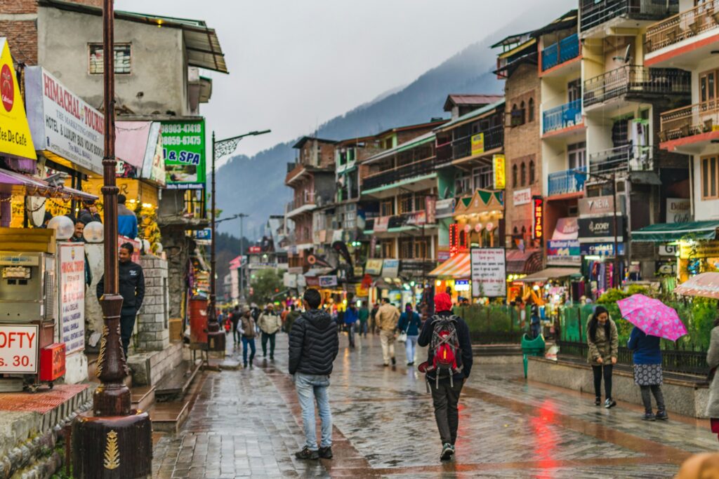 Crowded Manali street with roadside shops, hotels, cafes, and vehicles in a popular tourist area.
