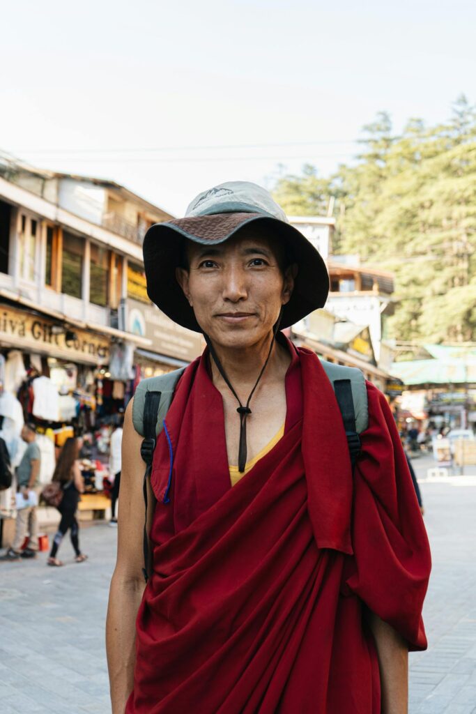 Buddhist monk standing on the colorful street leading to the Dalai Lama Temple in McLeod Ganj, surrounded by Tibetan culture and mountain vibes, captured for Visit Taxi travel and taxi services in Dharamshala, Amritsar, Chandigarh, Ludhiana, New Delhi and Jalandhar.