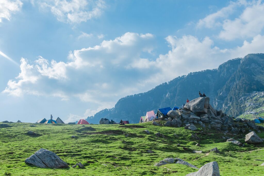 Panoramic view of the vibrant green, rolling mountains and rocky ridges along the Triund Trek trail under a clear sky.