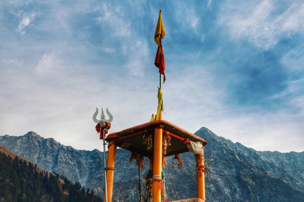 A small, stone Shiva temple adorned with colorful prayer flags, situated on a rocky mountain path along the Triund trek in Dharamshala, Himachal Pradesh, with the majestic Dhauladhar mountains in the background.