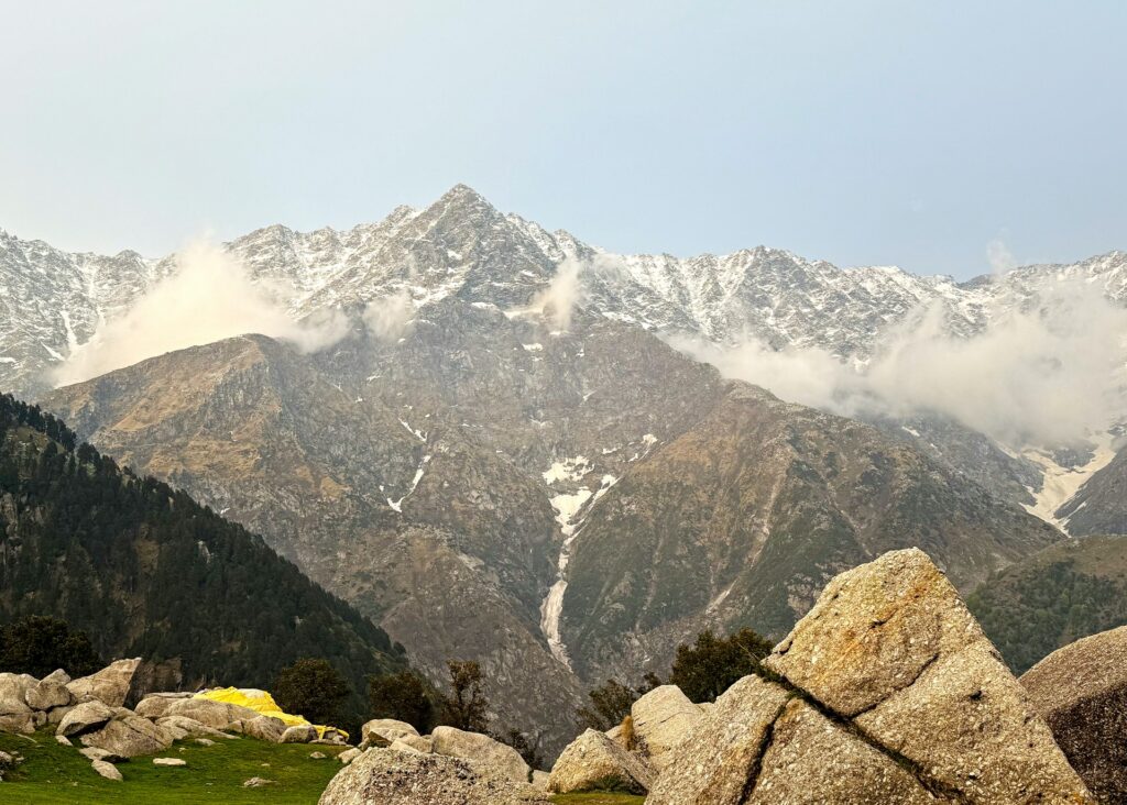 A scenic view of the Triund Trek near Dharamshala, featuring a contrast between rugged, rocky mountains and majestic snow-covered peaks under a clear sky.