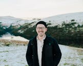 A young man wearing eyeglasses smiles while standing in a snow-covered outdoor setting.